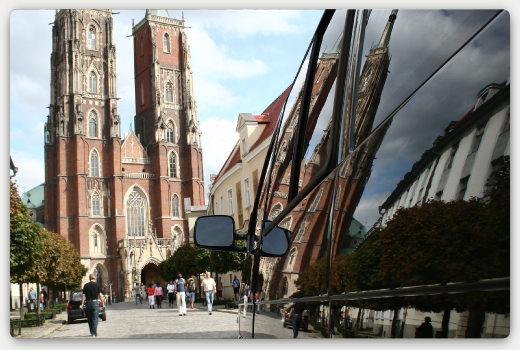 Odbicie Katedry we Wrocławiu w karoserii czarnego busa, widoczne lusterka boczne i spacerujący ludzie.